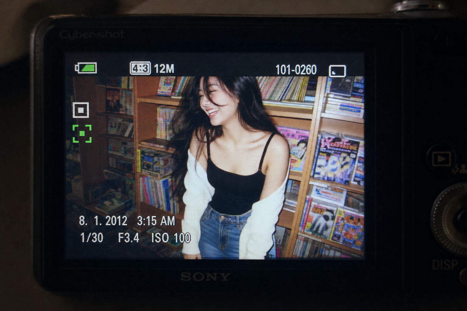 A glowing digital camera LCD screen showing a candid early-2010s flash photo of a young woman standing beside shelves of colorful manga and magazines, with retro camera UI overlays visible.
